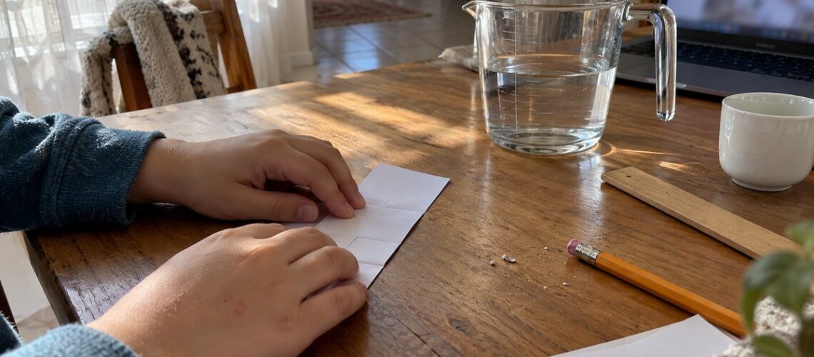 Close-up of a child's hands folding a paper strip beside a glass measuring jug on a wooden study table by a window.